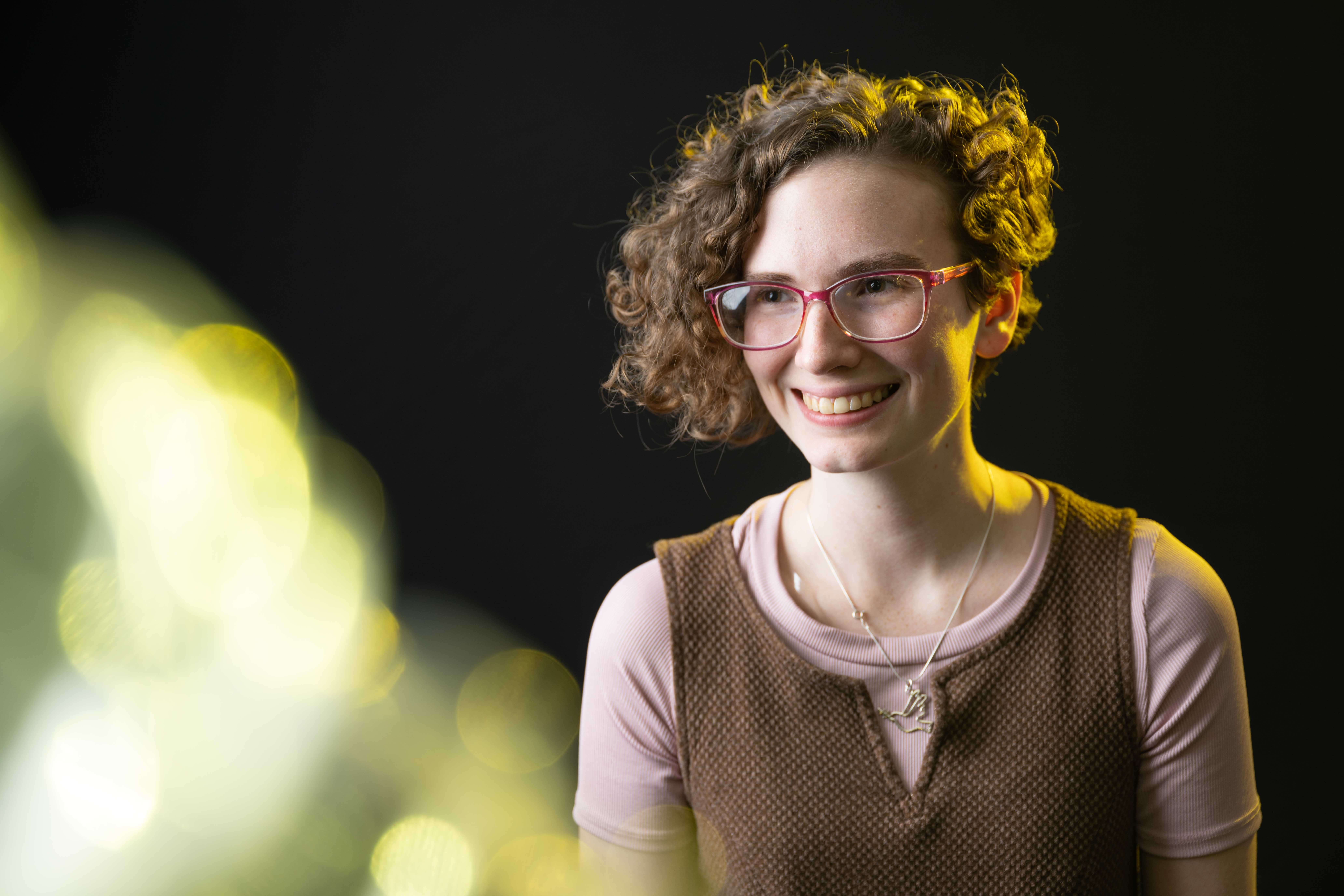 Flannery standing in front of a black background and smiling. She has short curly hair and is wearing glasses and a light purple and brown dress.